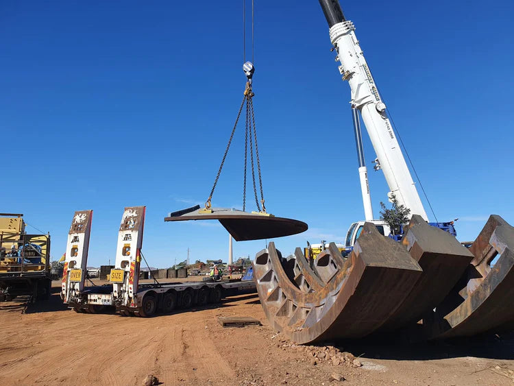 A large industrial piece of equipment, possibly a crawler track or heavy machinery component, suspended by a crane over a flatbed trailer on a dirt lot under a clear blue sky.
