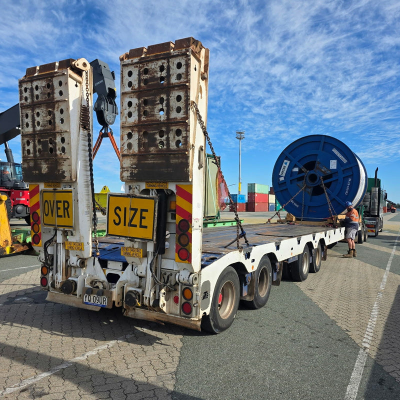 Truck carrying large cylindrical objects and a cable reel on a road with a blue sky.