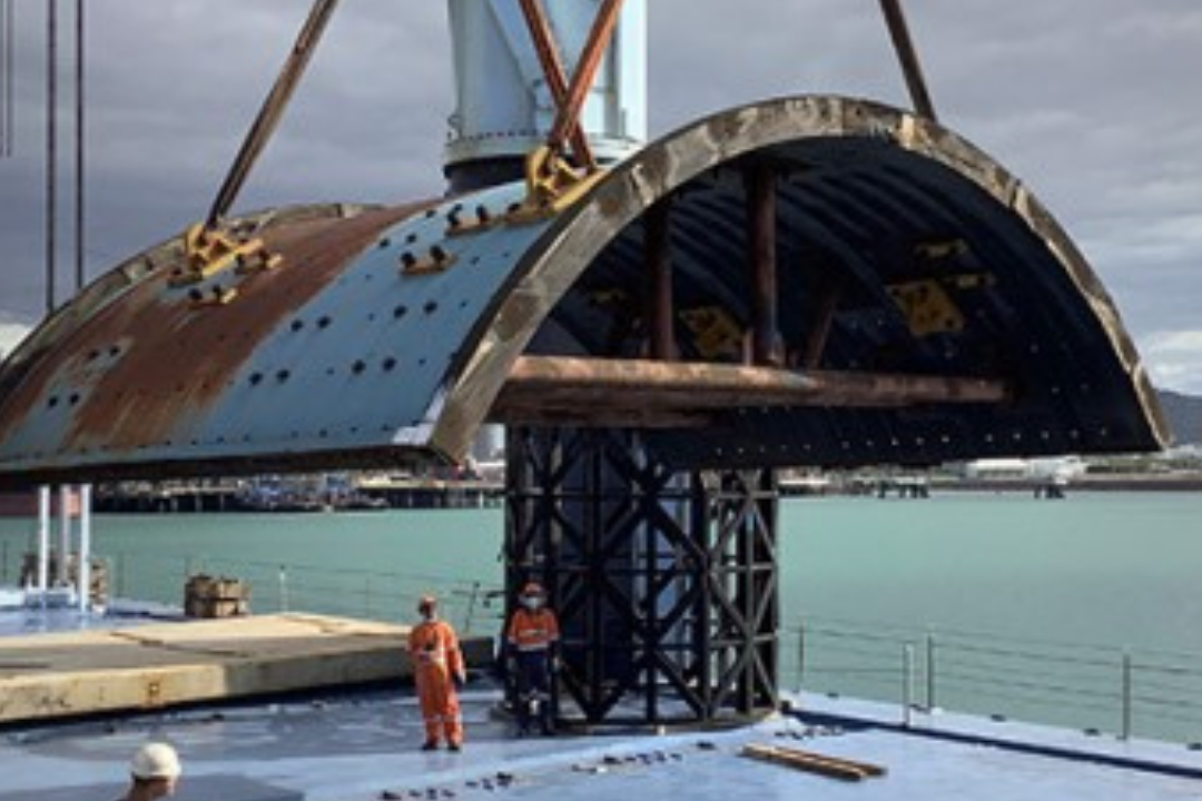 A large curved steel structure being lifted by a crane on a ship deck, with workers visible in the waterway setting under an overcast sky.